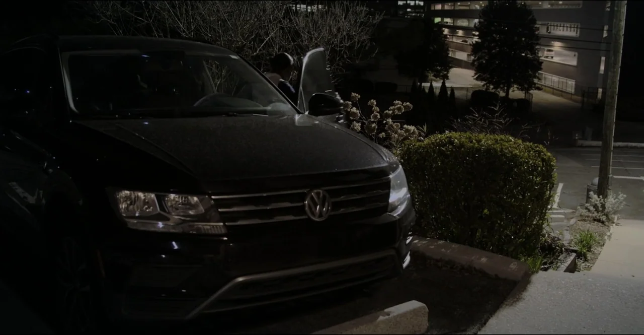 The Devil Is Busy (2024) movie still: medium — A dark SUV is parked in a suburban lot near a manicured hedge. A lone person stands by the open car…