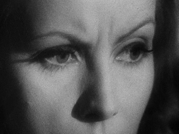 Queen Christina (1933) movie still: closeup — A close-up shot of a woman's face fills the frame against a dark background. She stares forward with a…