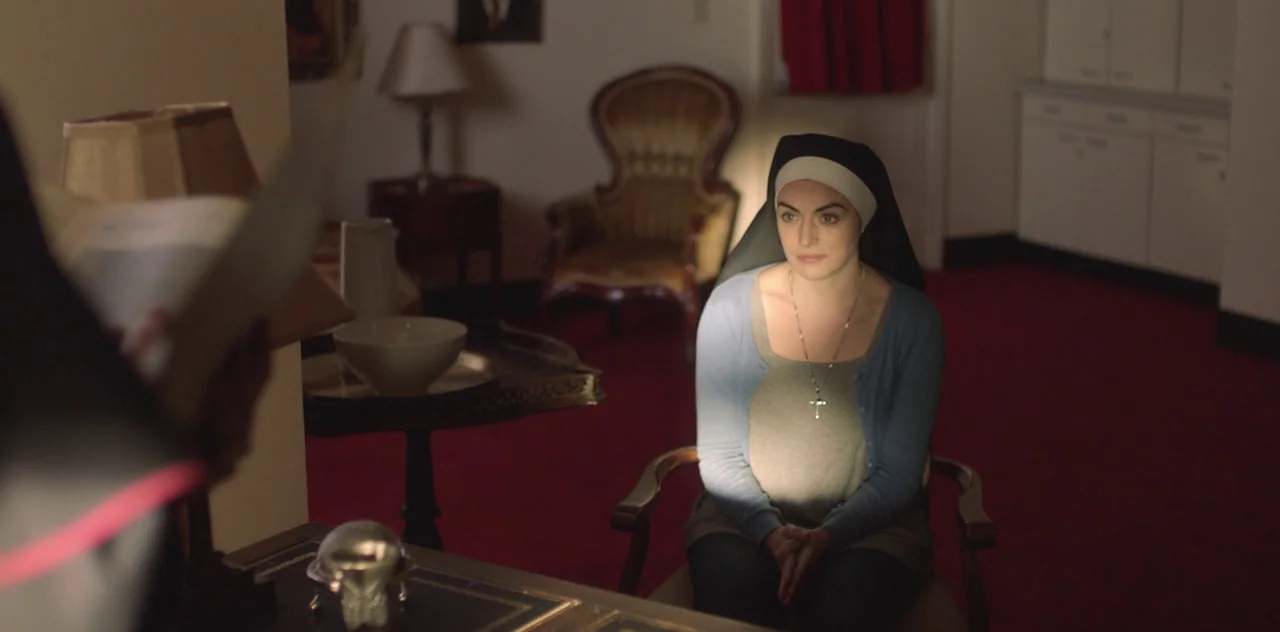 Darlin’ (2019) movie still: medium — An office with a red carpet floor features a desk, a lamp, and seating. A young woman dressed as a nun sits at…