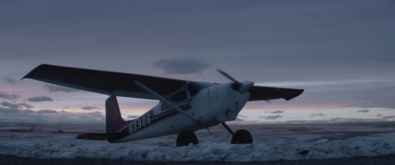 Hold The Dark (2018) movie still: wide — A small white and red private airplane rests on a snow-covered field. Distant mountains are visible under a…