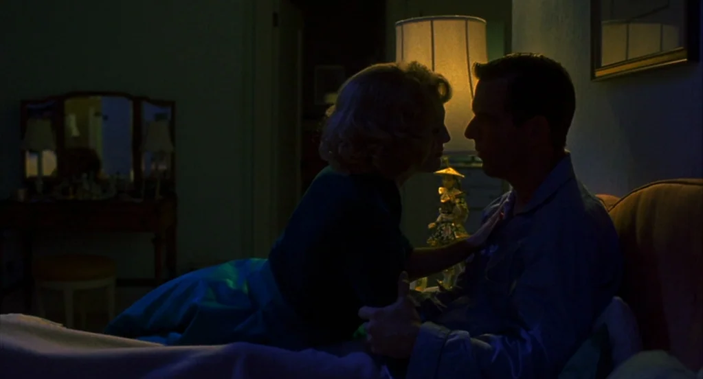 Far From Heaven (2002) movie still: medium — A dimly lit room at night with a bedside lamp emitting soft golden light. A woman in a dark dress leans…