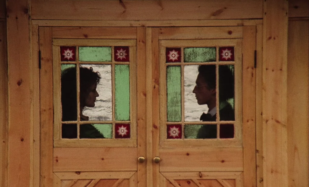 Mahler (1974) movie still: medium — A light wood double door features two square windows with decorative green and red stained glass. A woman and a…