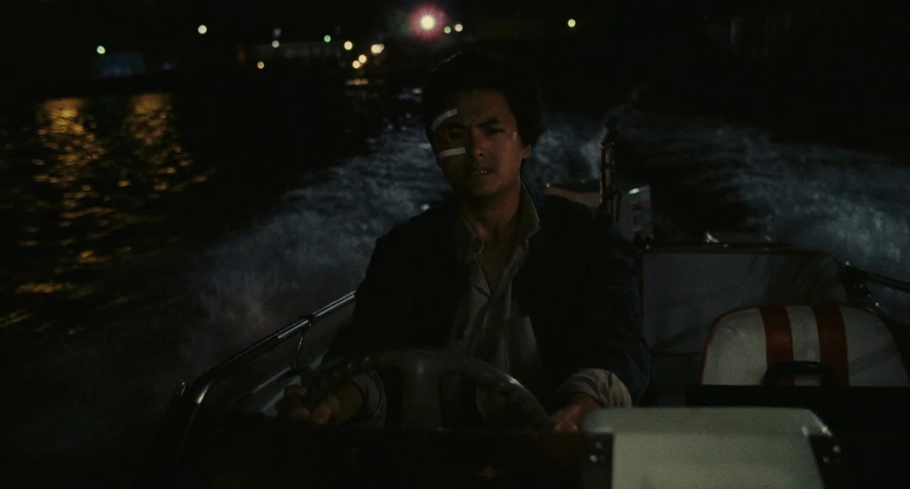 A Better Tomorrow (1986) movie still: medium — A man drives a small motorboat through dark water under the night sky. City lights twinkle in the…
