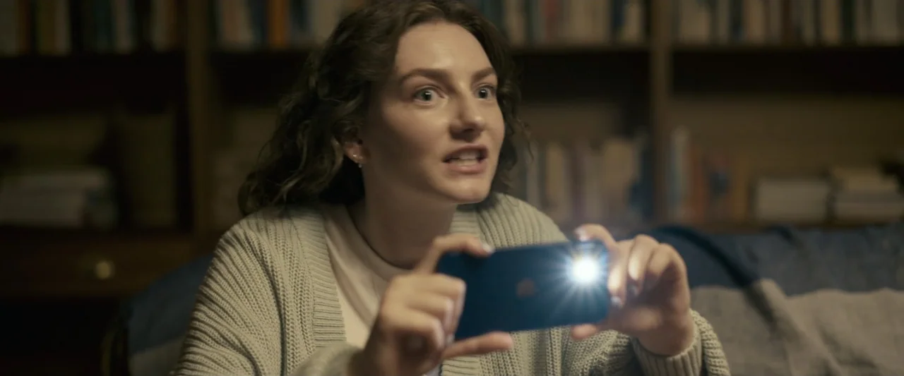 Talk to Me (2023) movie still: medium — A dimly lit room contains a bookshelf in the background and soft textures. A young woman holds a smartphone…