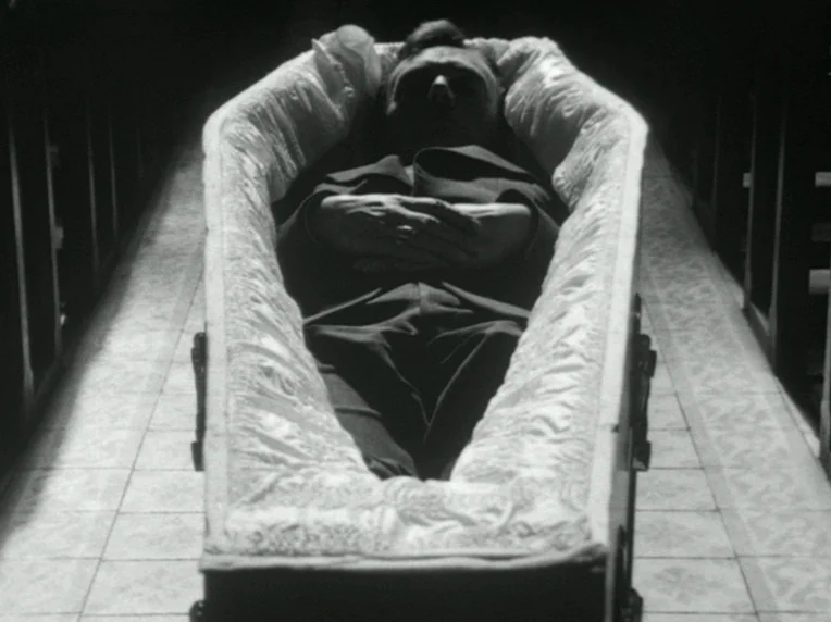 Death and Transfiguration (1983) movie still: medium — A man lies inside a lined coffin situated in the center of an aisle. The dark space is…
