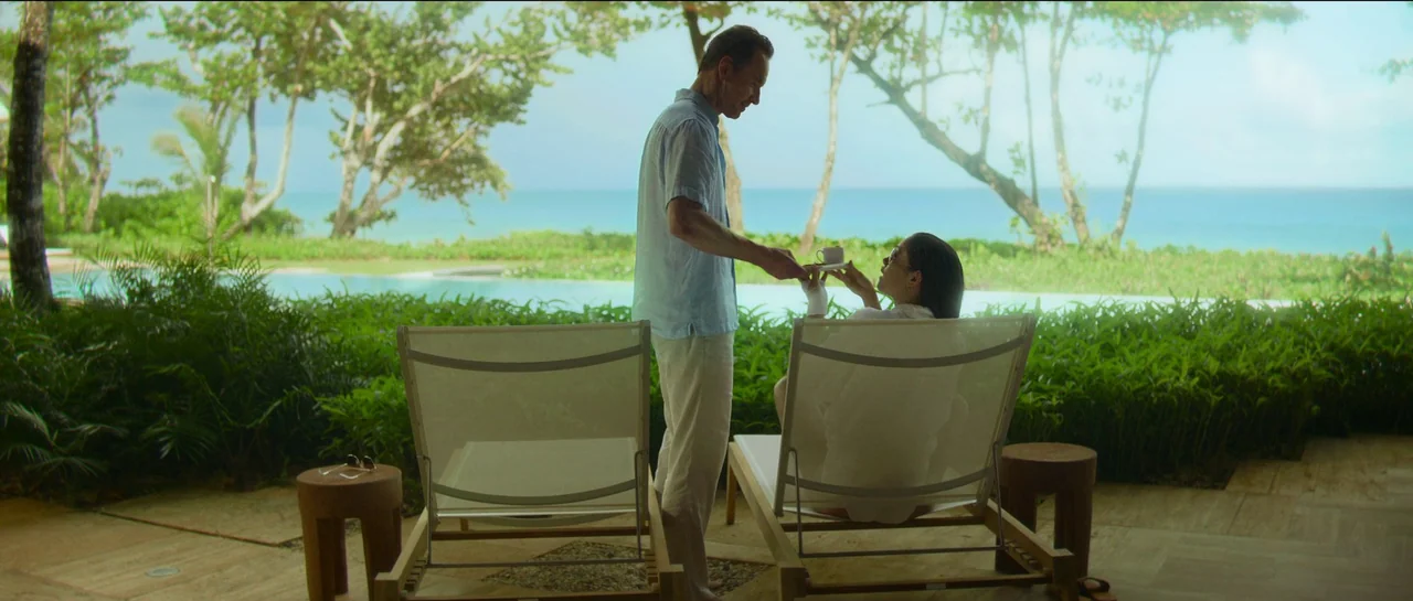 The Killer (2023) movie still: fullbody — An open-air resort patio features two lounge chairs facing a turquoise ocean and pool. A man in a blue…