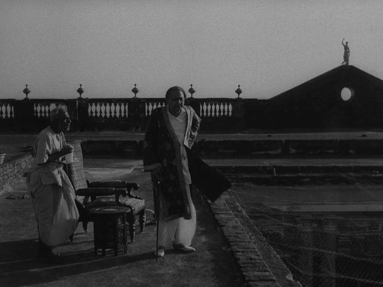 The Music Room (1958) movie still: wide — A spacious stone terrace with a decorative balustrade overlooking a building roofline. Two elderly men…