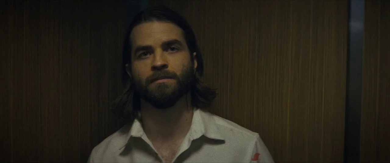 Synchronic (2019) movie still: closeup — A confined space lined with vertically grained wood paneling in muted brown tones. A man with a full beard…
