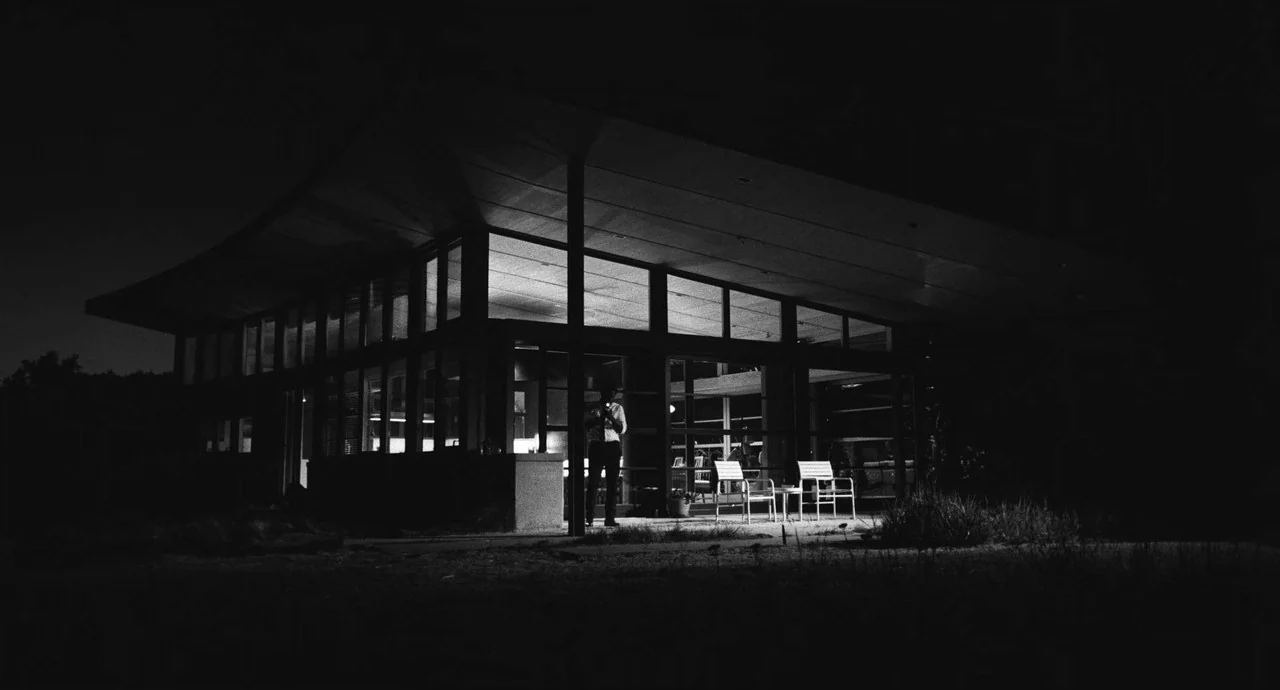 Malcolm & Marie (2021) movie still: wide — A modern building with floor-to-ceiling windows is illuminated from within at night. The surrounding area…
