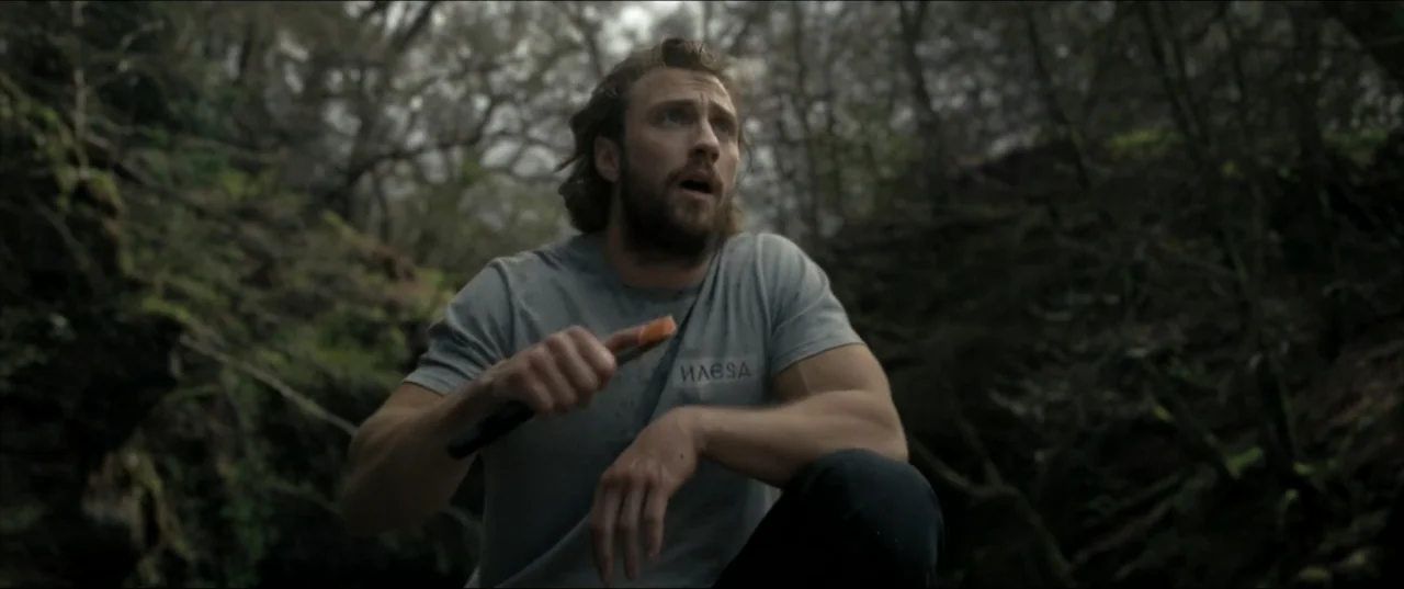 Kraven the Hunter (2024) movie still: medium — A dense, overgrown ravine with tangled trees and shadows in the background. A bearded man crouches in…