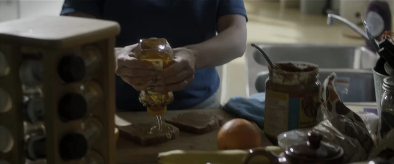The Hollow Child (2018) movie still: medium — A cluttered kitchen counter surface contains food items and a spice rack. A person squeezes golden…