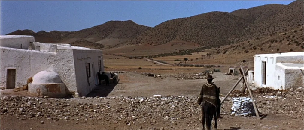 Fistful Of Dollars (1964) movie still: wide — A remote desert settlement features weathered white stucco buildings with a rocky ground surface. A…