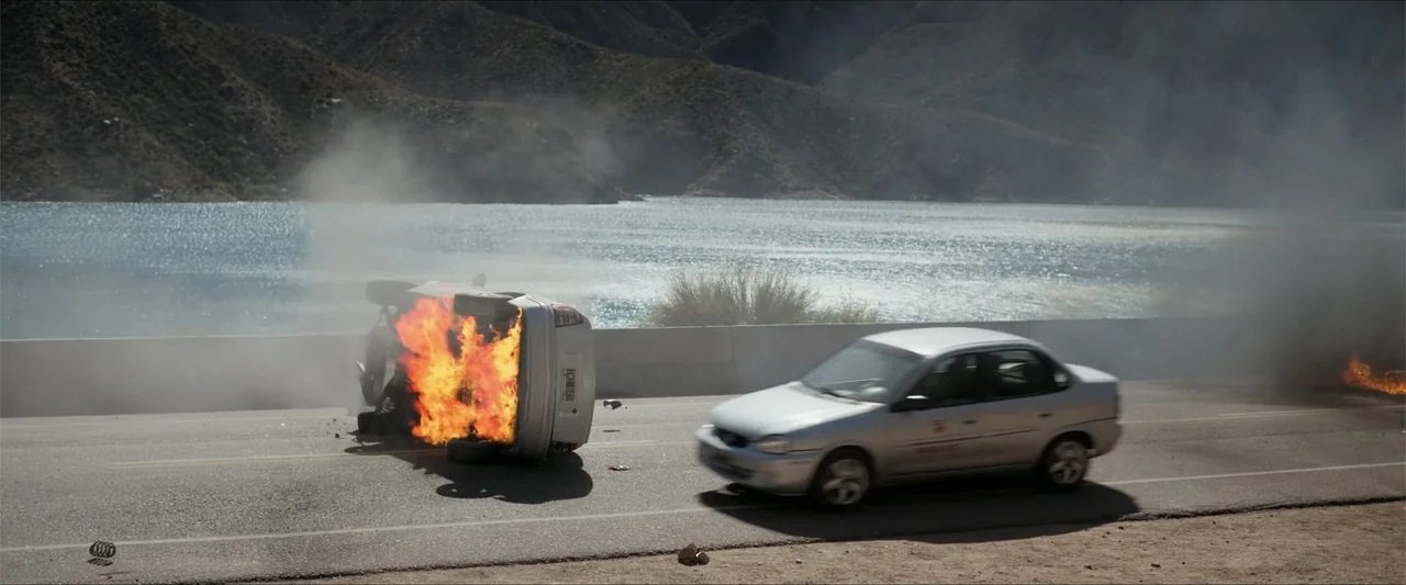 Checkmate (2015) movie still: wide — A silver car drives along a lakeside road next to an overturned burning vehicle. Steep mountains rise in the…
