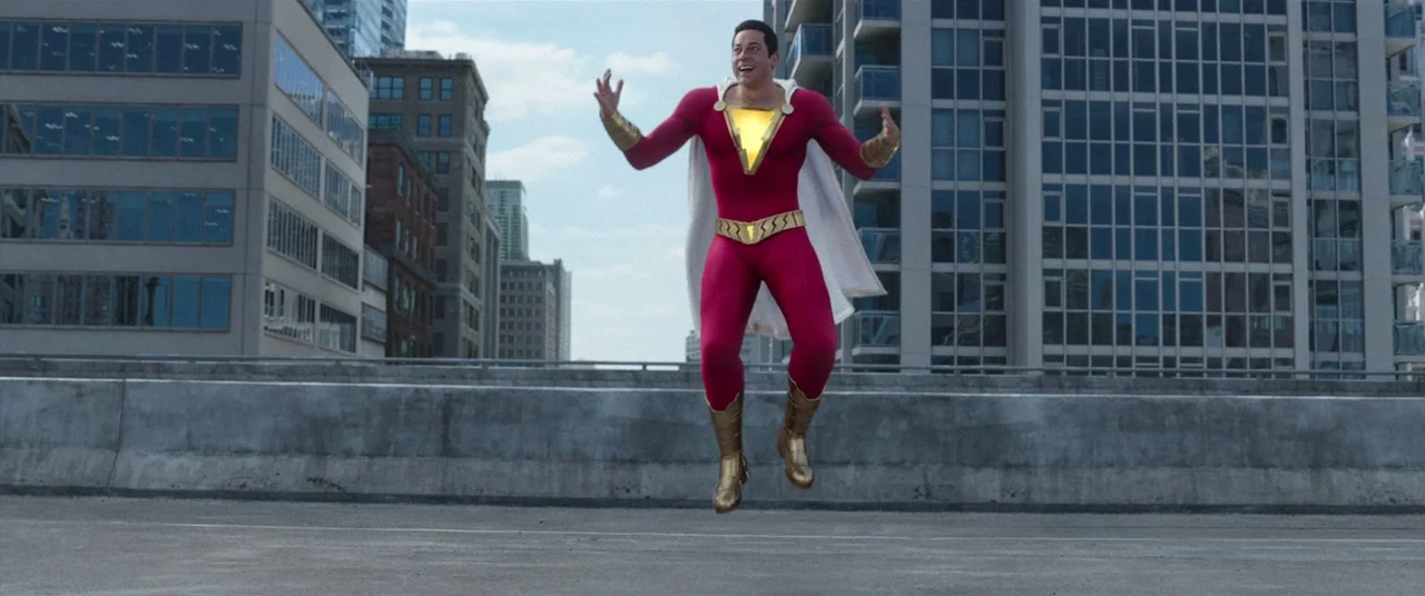 Shazam! (2019) movie still: fullbody — A man in a bright superhero costume hovers in the air above a highway. Modern high-rise glass apartment…