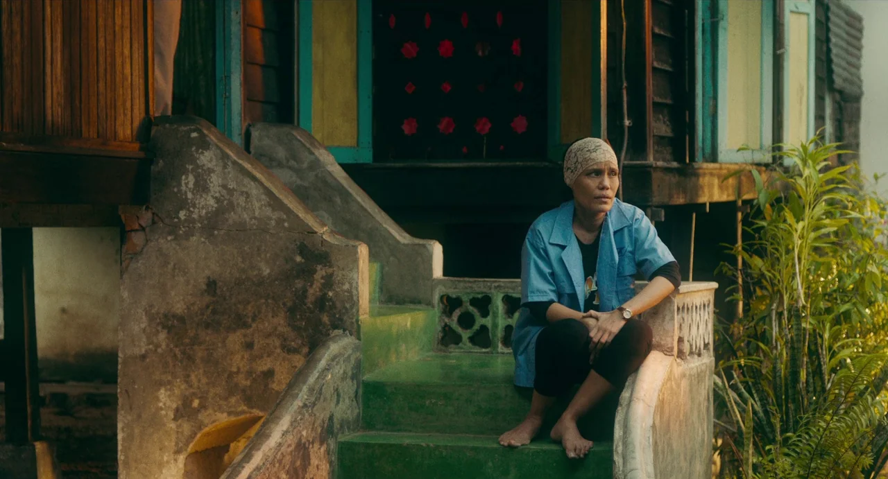 Tiger Stripes (2023) movie still: medium — A woman sits on the concrete steps of a home with colorful painted architectural details. She rests on…