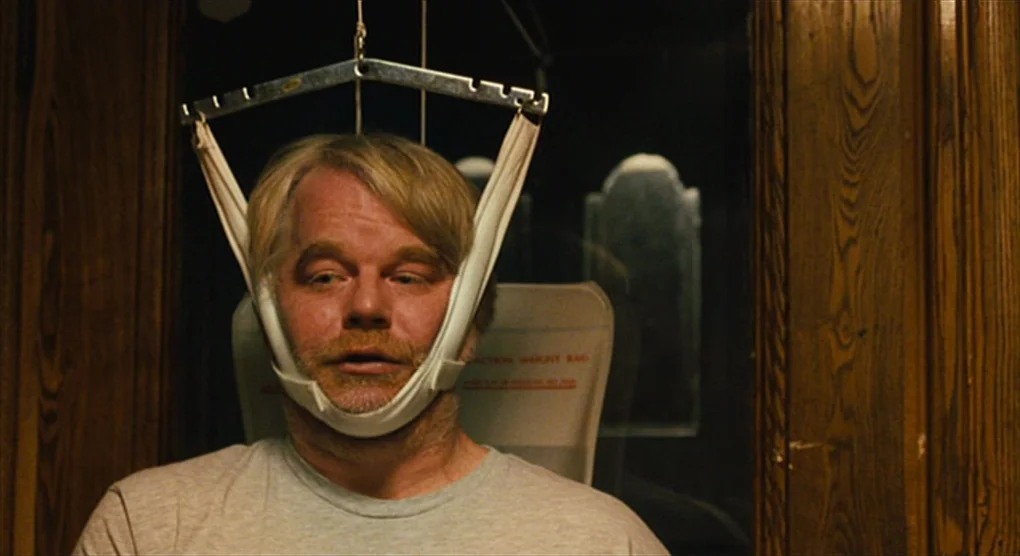 The Savages (2007) movie still: medium — A man stands before a wood-paneled wall with a dark reflective surface. He wears a medical cervical…