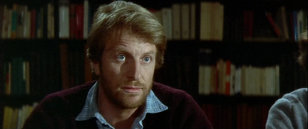 Love Unto Death (1984) movie still: closeup — A man sits in front of a dark, packed bookshelf in a room. He looks towards the left of the frame…