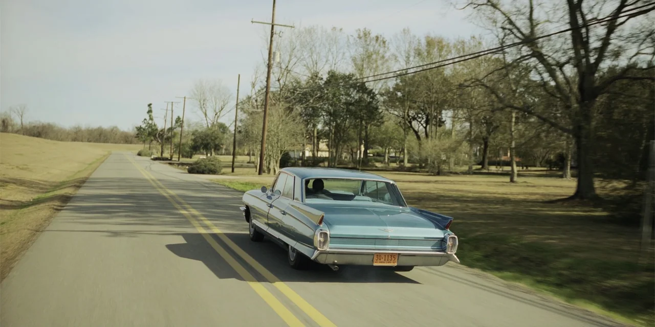 Green Book (2018) movie still: wide — A rural asphalt road cuts through a landscape with sparse trees and dry grass. A vintage teal-colored Cadillac…