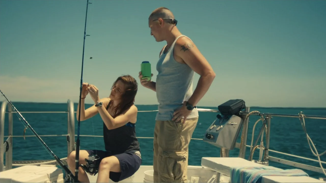 Camp X-Ray (2014) movie still: medium — The deck of a boat is surrounded by open blue ocean under clear skies. A woman fixes her fishing line while…