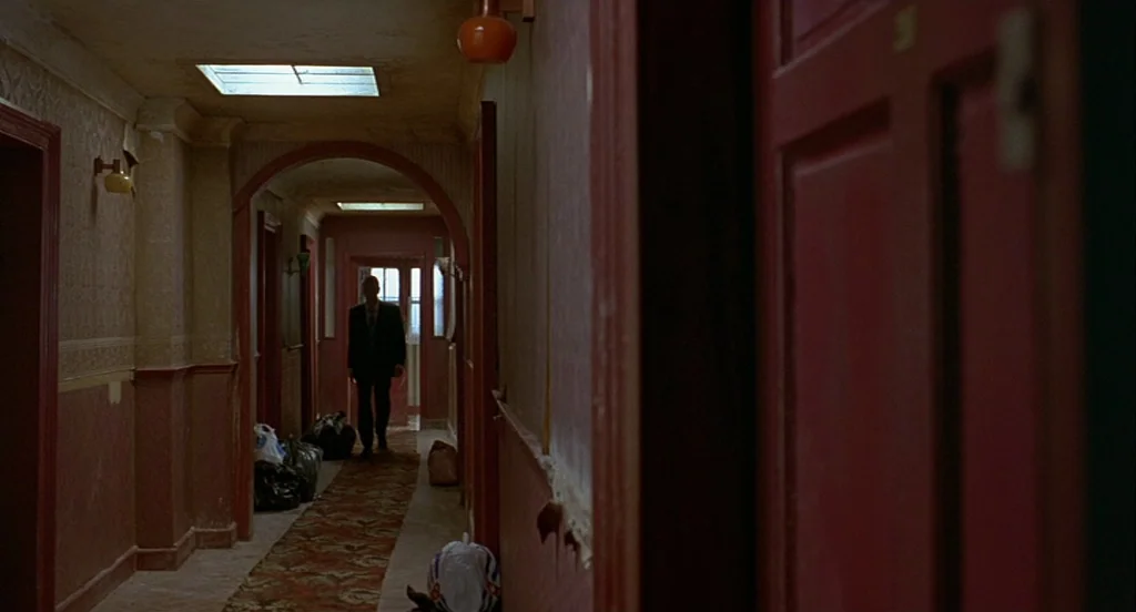 Trainspotting (1996) movie still: wide — An aging apartment hallway features patterned wallpaper, a long worn runner rug, and multiple doors. A man…