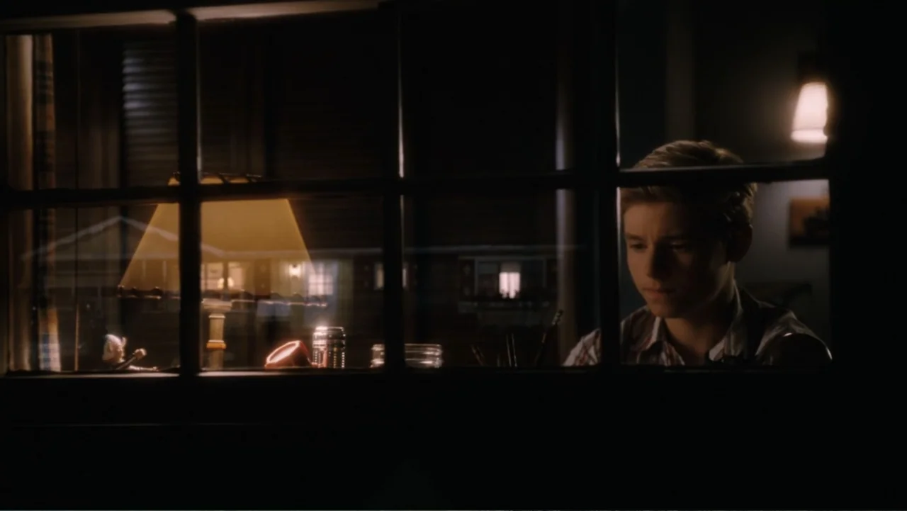Flipped (2010) movie still: medium — A dark interior room features a wooden desk with a lit lamp at night. A young boy sits at the desk looking down…