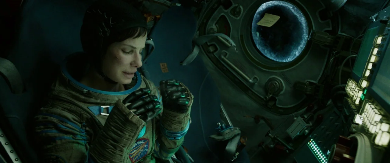Gravity (2013) movie still: medium — A cramped spacecraft cockpit with complex instrument panels and a circular window. A woman in a flight suit…