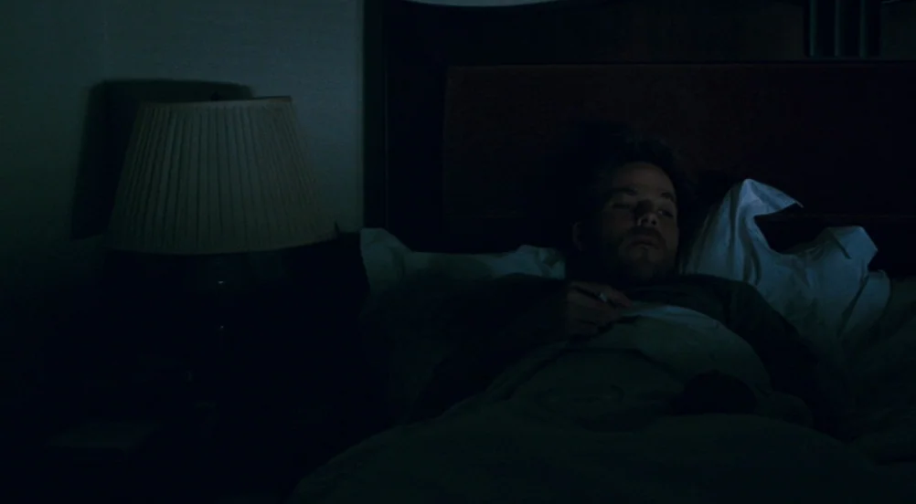 Somewhere (2010) movie still: medium — A darkened room features a man lying under white sheets on a bed frame. The man rests in the dim light of an…
