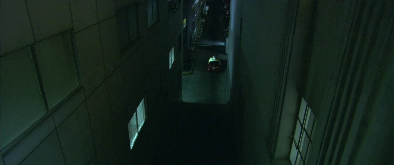 Collateral (2004) movie still: establishing — A narrow concrete alleyway between two tall windowed buildings at night. A lone taxi is parked on the…