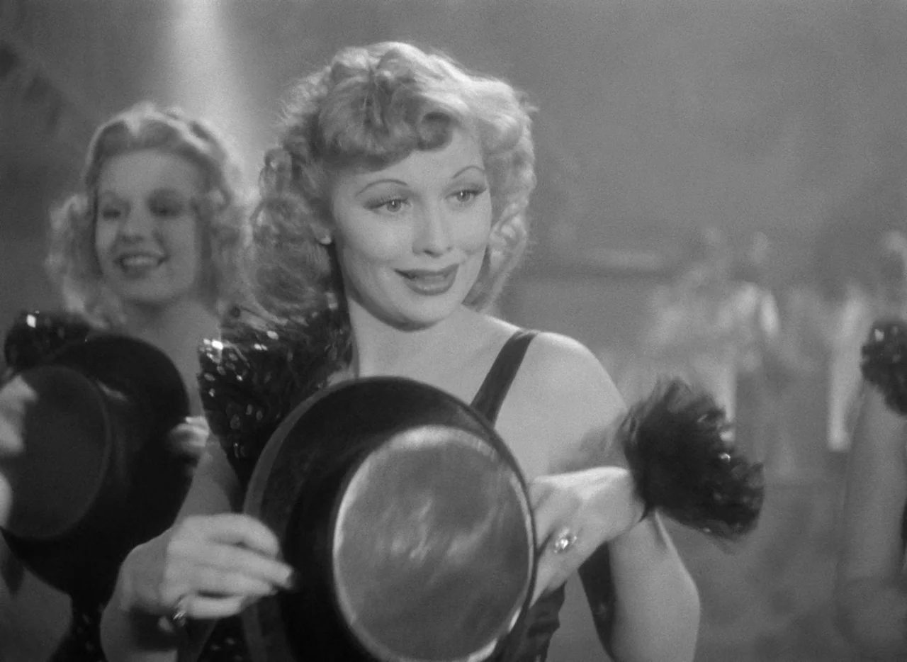 Dance, Girl, Dance (1940) movie still: medium — A brightly lit performance space with blurred figures in the background. Two women stand in costumes…