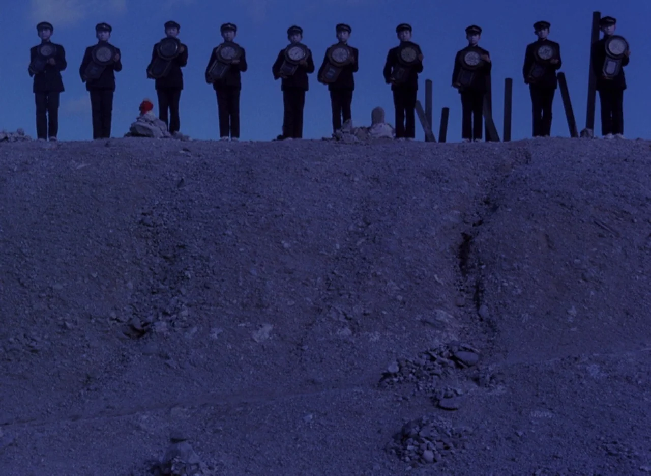 Pastoral To Die In The Country (1974) movie still: wide — A line of ten children in dark uniforms stands atop a barren gravel embankment. Each child…