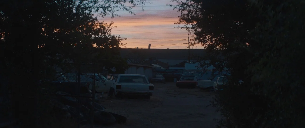 Honey Boy (2019) movie still: establishing — A dark salvage yard contains multiple rusted vintage automobiles parked on a dirt lot. Trees frame the…