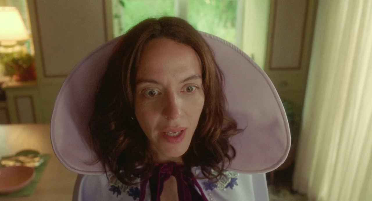 Babysitter (2022) movie still: closeup — An interior space features a person wearing a formal, retro-style lavender cape. The subject is centered in…