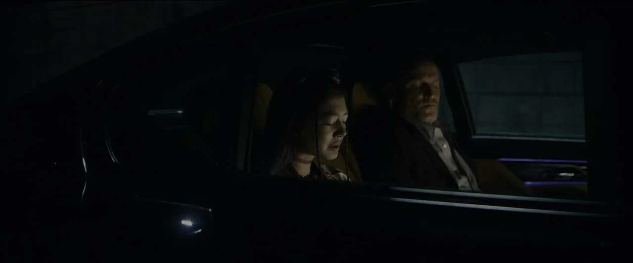 Kate (2021) movie still: medium — The interior of a modern car at night with two people sitting in the backseat. A young woman and an older man sit…