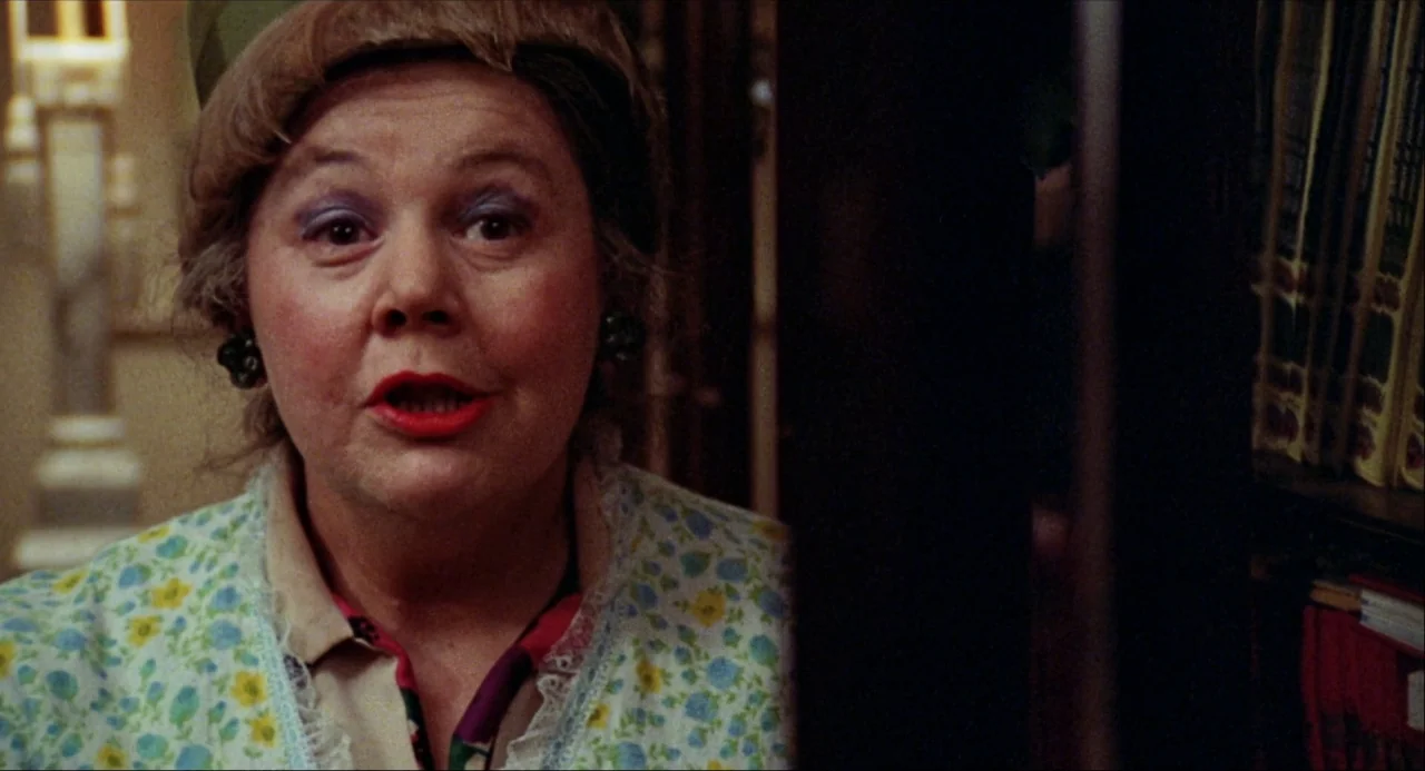 Black Christmas (1974) movie still: closeup — A woman stands in a dim room with a bookshelf visible on the side. She looks toward the camera with…