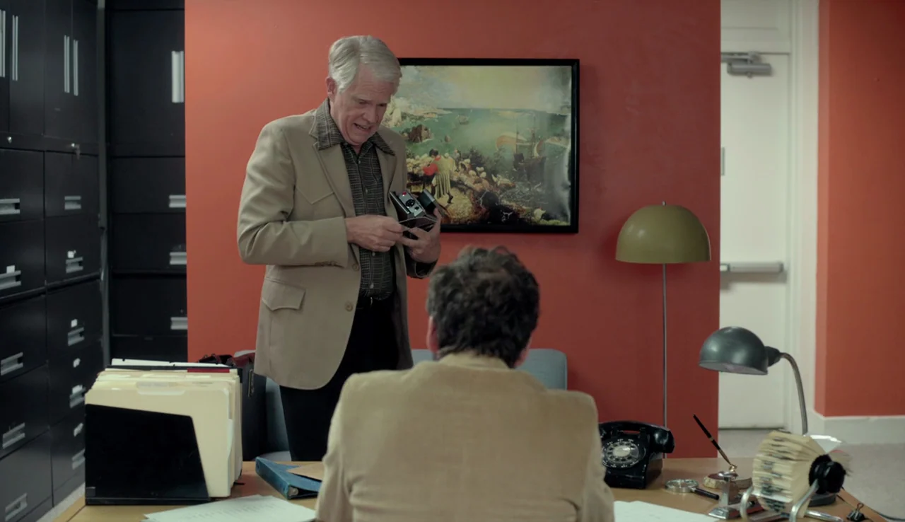 Experimenter (2015) movie still: medium — An office features a vibrant orange wall and rows of black metal filing cabinets. A man presents a folding…