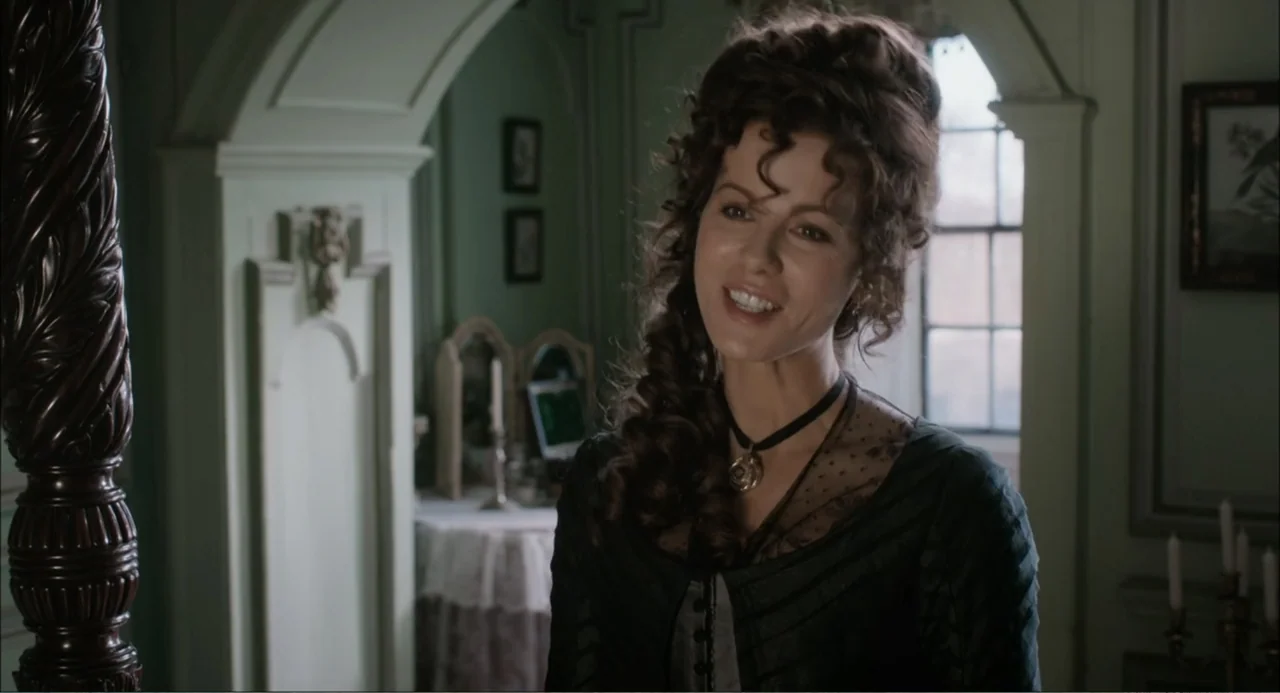 Love & Friendship (2016) movie still: medium — A light-walled room features an arched doorway and a mahogany bedpost. A woman in a Victorian-style…