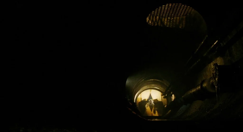 Mimic (1997) movie still: wide — A dark, cylindrical concrete sewer tunnel recedes into a distant illuminated opening. A circular metal grate sits…