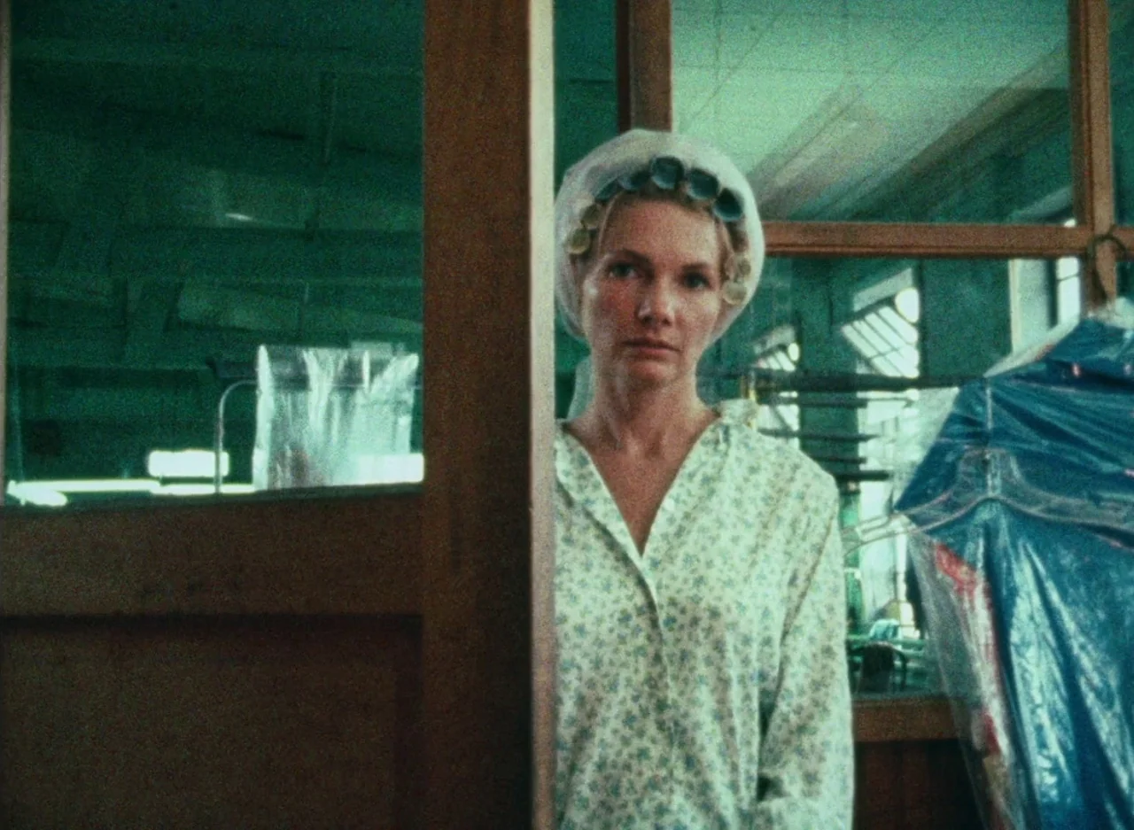Wanda (1970) movie still: medium — A utilitarian space with wooden framing dividers and racks of plastic-covered garments. A woman with hair curlers…