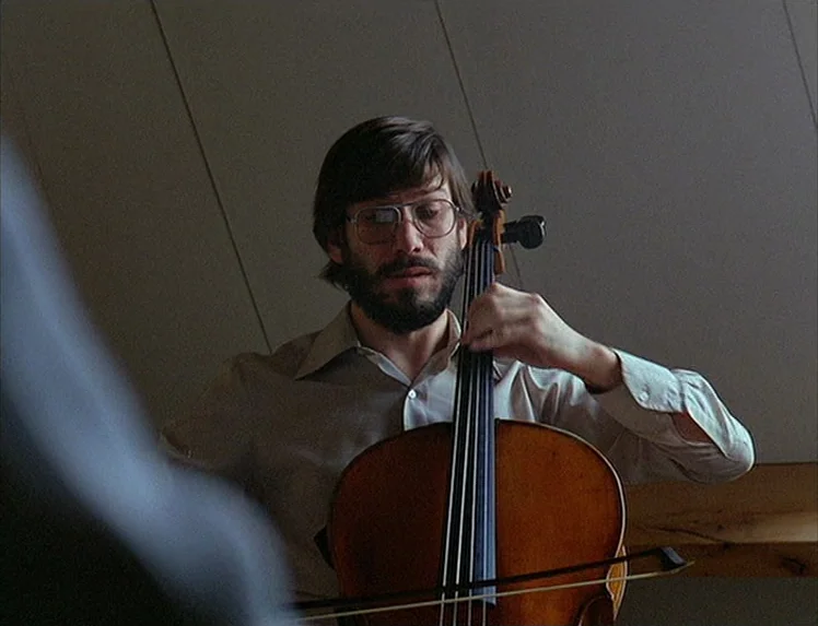 First Name: Carmen (1983) movie still: medium — A man plays a cello inside a room with plain walls. He focuses on the strings while holding the bow…