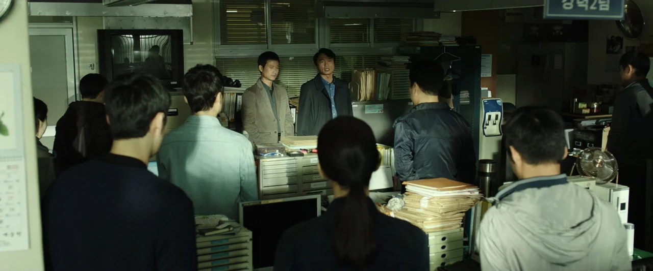 The Gangster, the Cop, the Devil (2019) movie still: medium — A cluttered police office with filing cabinets, stacks of paper, and venetian blinds.…