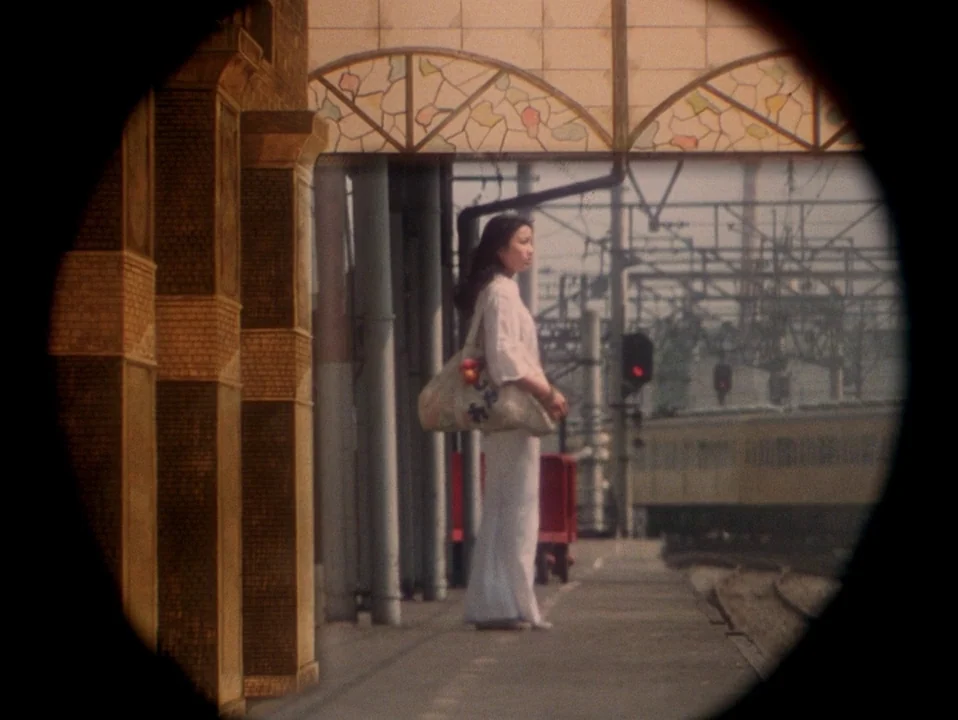 House (Hausu) (1977) movie still: fullbody — A woman stands on an outdoor railway platform with arched glass roofing and brick pillars. She holds a…