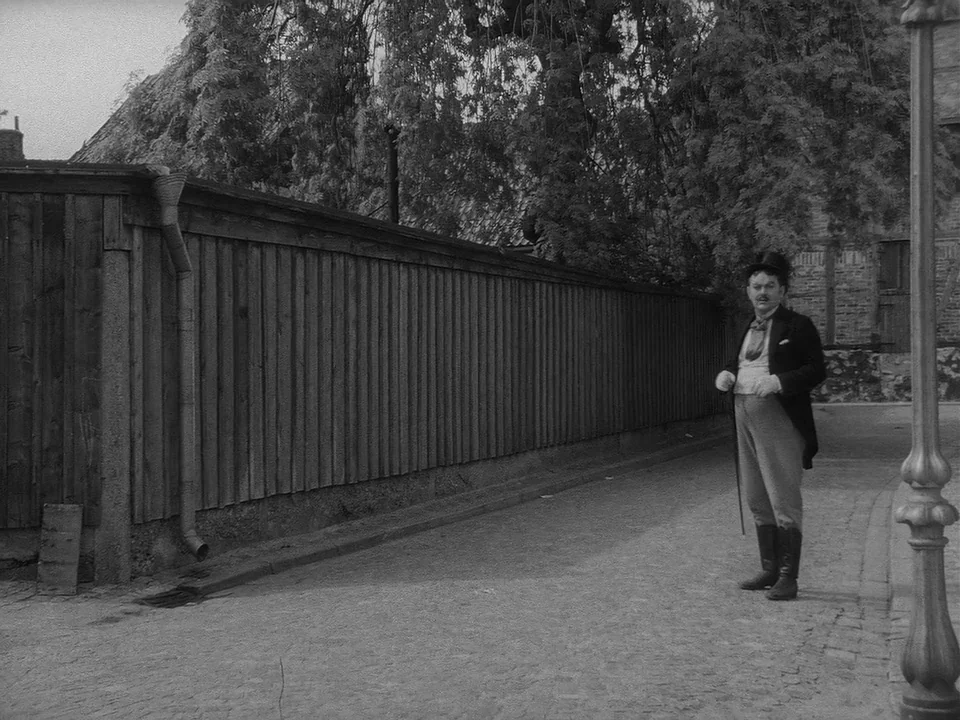 Sawdust and Tinsel (1953) movie still: fullbody — A narrow cobblestone street is bordered by a long vertical wooden plank fence. A man in formal…