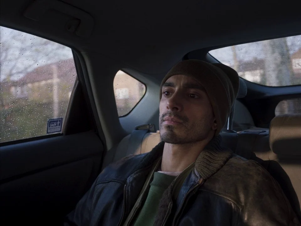 Mogul Mowgli (2020) movie still: medium — A man sits in the back of a car during a rainy day. Raindrops streak the window beside him as he gazes…