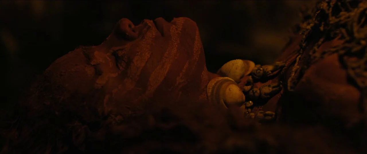 Out of Darkness (2022) movie still: closeup — A dimly lit cave space surrounds a figure lying in the darkness. The person is resting with clay…