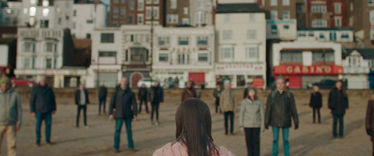 Saint Maud (2019) movie still: medium — A group of people stands on a beach in front of a line of buildings. A woman in a pink jacket faces the…