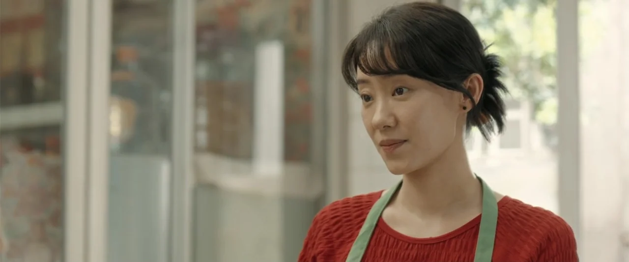 Yolo (2024) movie still: medium — A well-lit shop interior features a counter with glass panels and a blurred background. A young woman in a red…