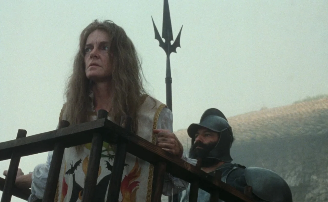 The Pit and The Pendulum (1991) movie still: medium — A woman and an armed guard stand on a wooden platform against a grey sky. A metal-tipped pole…