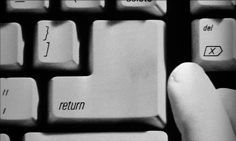 Pi (1998) movie still: closeup — A close-up of a vintage computer keyboard with a large return key. A human finger is visible resting near the…