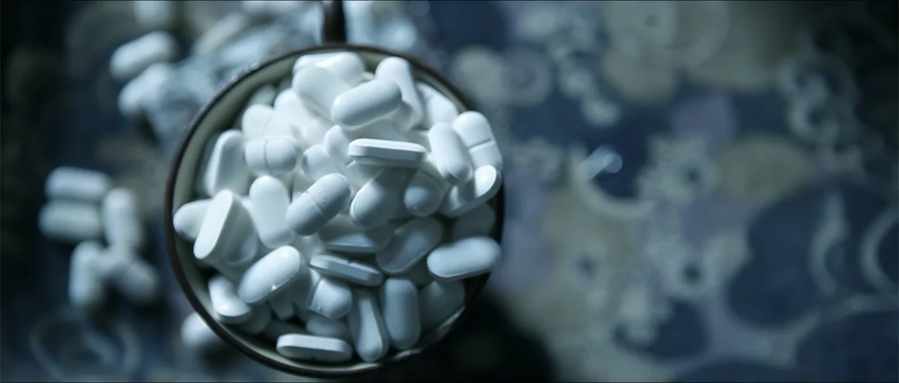 Garnet’s Gold (2014) movie still: closeup — A circular cup holds a dense mound of white pharmaceutical pills on a surface. Some individual pills lie…