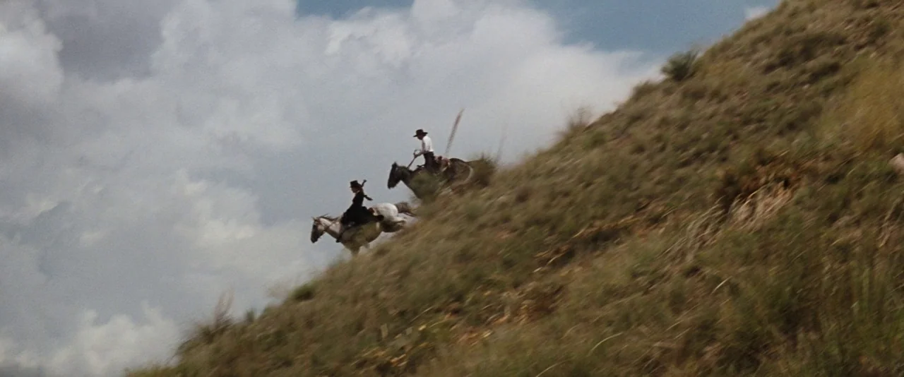 Tombstone (1993) movie still: wide — Two riders on horseback traverse a steep, dry grassy incline beneath an overcast sky. The pair moves…